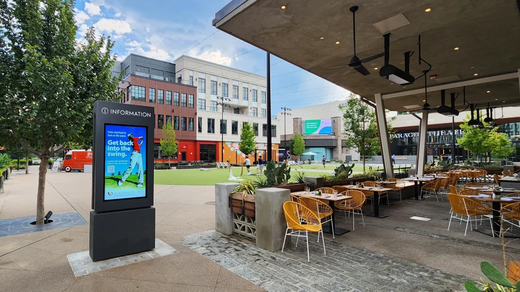 A patio next to an open grass square surrounded by shops with an outdoor LCD display