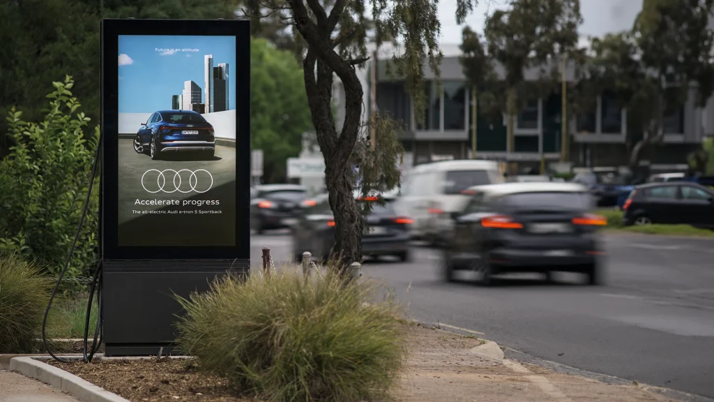 An outdoor display with EV charger shows an ad next to a busy street