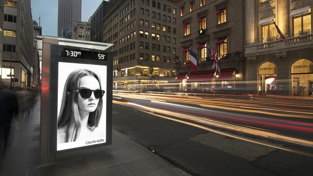 Advertisement of a woman wearing sunglasses - black and white - on a large digital screen attached to bus shelter in a city