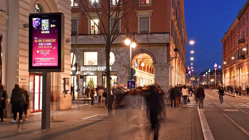 A bright and colorful display on a post next to a number of people walking down a well lit city street