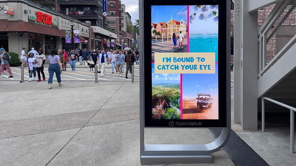 A street-side digital kiosk displays a colorful travel collage with pedestrians nearby