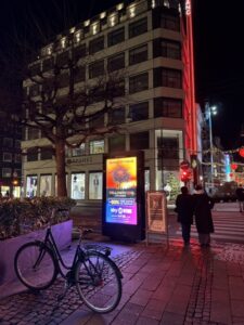 A city street with an office building in the background at night with a bright colorful digital display and a bicycle in the foreground