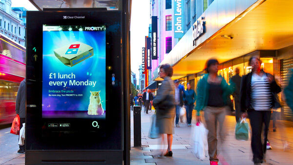 A sidewalk digital advertising kiosk shows a colorful promo with people walking by