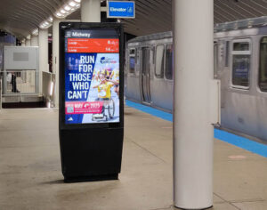 A digital display in a subway with a row of columns and a train in the background