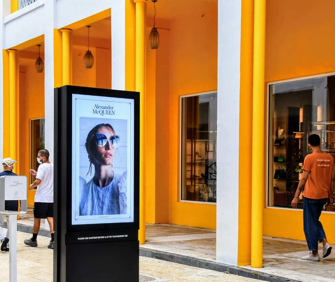 A retail building with columns painted bright orange and white in front of a street with pedestrians a digital display running an ad