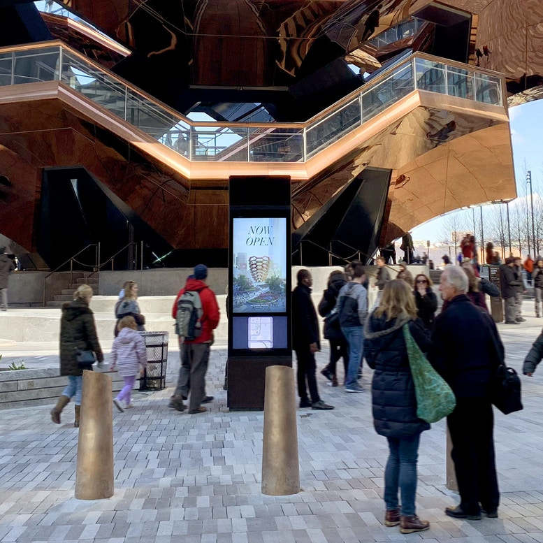 A large, artistic building covered in copper behind a crowd of pedestrians and a digital outdoor display