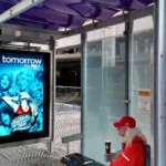 A person in a red outfit sitting in a bus shelter next to a large digital display