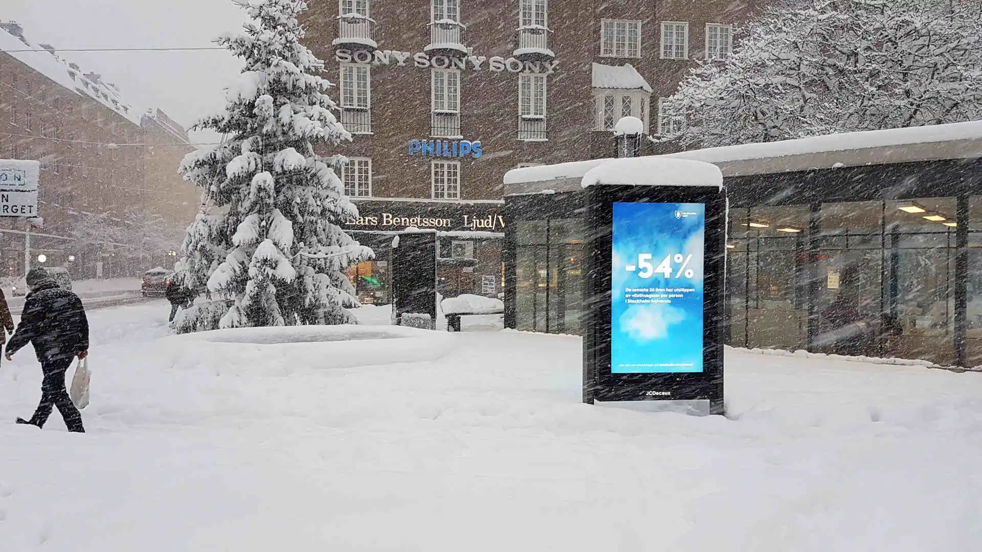 A digital display in front of a glass window wall in the middle of town which is covered with a thick layer of snow