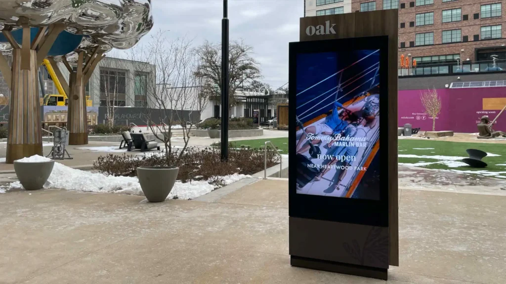An LCD display in front of a common area with shops surrounding and light snow on the ground