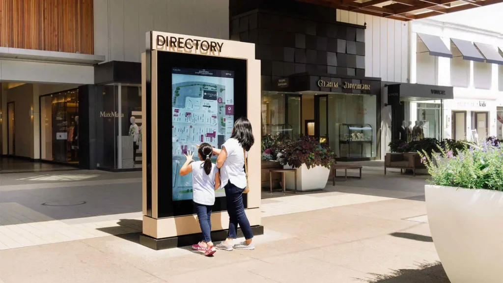 A brightly lit outdoor retail strip with a directory display being used by two girls