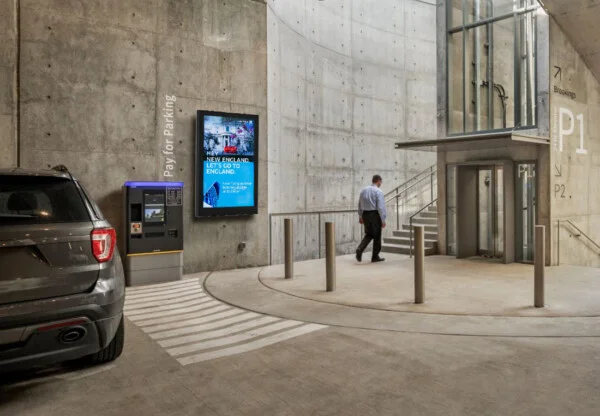 a man walking away from a gray cement parking garage towards a staircase-elevator, there is a wall mounted LCD with an ad