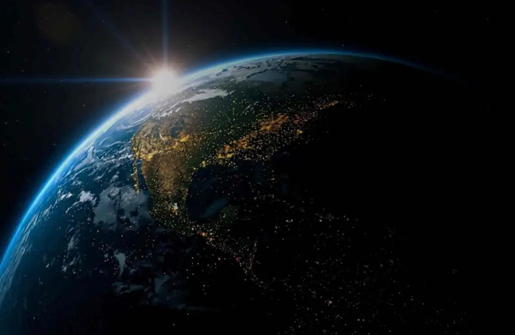 A view of the planet but more of a silhouette with the sun peaking over the horizon and little dots showing city lights