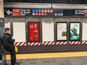 A subway train station with a platform and passenger in front of 3 digital displays and directional signage above