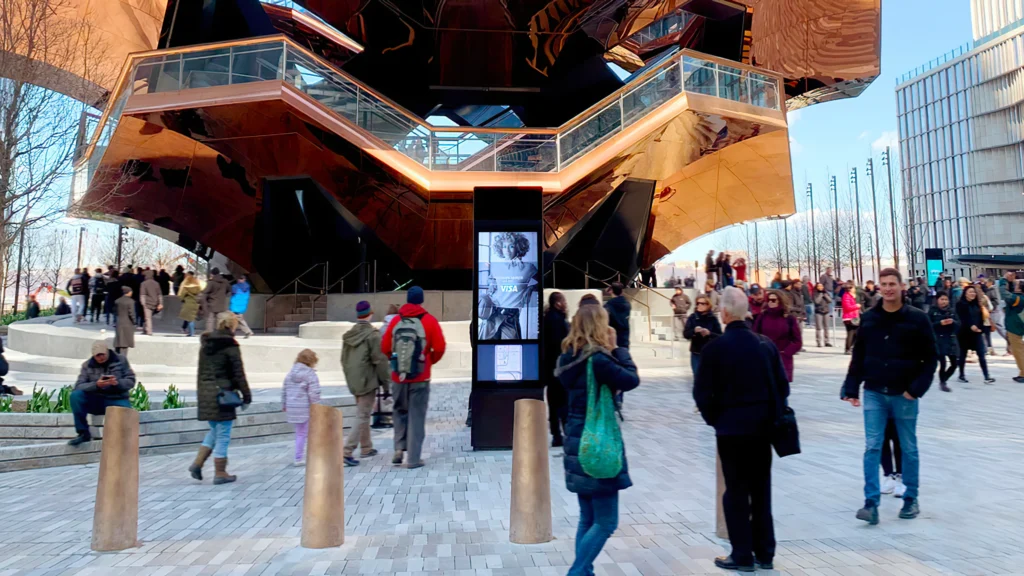 A large copper building with obscure architecture surrounded by people walking in a pave pavilion.